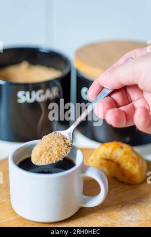Espresso appena versato, caldo e pronto da bere a colazione, un cucchiaio d'argento che sta per ammorbidire lo zucchero di canna, su una tavola di legno, un panino, zucchero Foto Stock