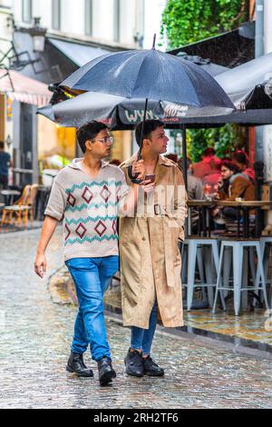 Due amici uomini che camminano per strada sotto un ombrello durante il temporale - Bruxelles, Belgio. Foto Stock