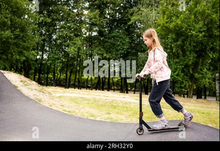 Preteen girl che guida uno scooter nel parco. Carina bambina con veicolo ecologico all'aperto Foto Stock