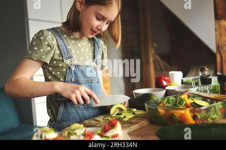 La bambina taglia l'avicado con il coltello in cucina. Bella bambina con frutta e verdura che prepara l'insalata Foto Stock