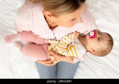 Mamma premurosa che tiene in mano la figlia piccola, bambina addormentata con il succhietto in bocca, sopra la vista Foto Stock