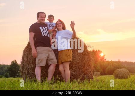 La famiglia sorride calorosamente mentre tiene il suo bambino in mezzo alla bellezza della natura, in piedi su un campo punteggiato da pile di fieno sullo sfondo di un'ora Foto Stock