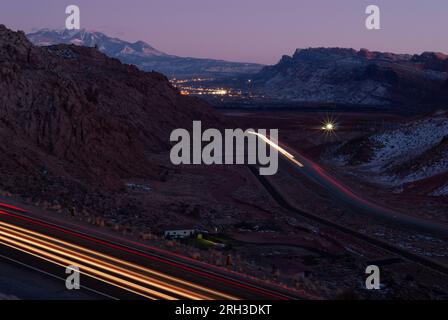 Le luci delle auto e la valle del fiume Colorado dopo il tramonto, con la città di Moab sullo sfondo. Vicino all'ingresso dell'Arches National Park, Utah, Stati Uniti. Foto Stock