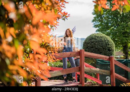 Splendido giardiniere allo zenzero che guarda qualcosa mentre si trova in piedi su un ponte di legno rosso nel giardino con un rastrello sulle spalle Foto Stock