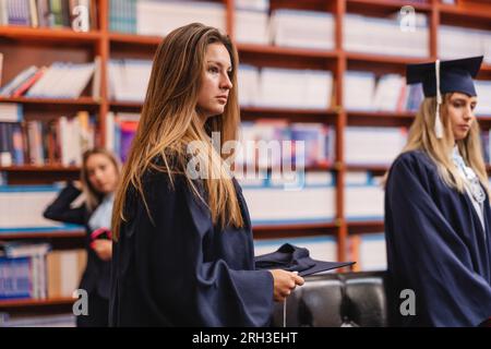 Giovane e attraente laureata in camera con i suoi colleghi pronti a scattare la foto della laurea in biblioteca Foto Stock