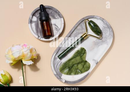 Flacone in vetro marrone con pipetta, massaggiatore manuale in pietra verde e raschietto per procedure cosmetiche su sfondo beige, vista dall'alto Foto Stock