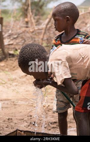 Giovani bambini kenioti che a turno bevono da una pipa d'acqua dolce con acqua proveniente dal vicino foro, contea di Baringo, Kenya Foto Stock