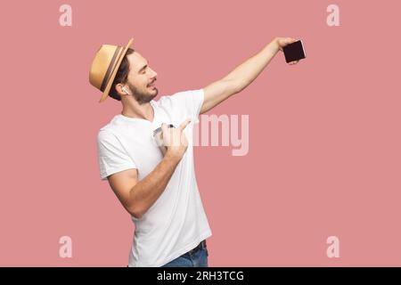 Ritratto dell'uomo blogger barbuto con T-shirt bianca e cappello in piedi con lo smartphone in mano e in diretta streaming, bevendo un caffè. Riprese in studio per interni isolate su sfondo rosa. Foto Stock