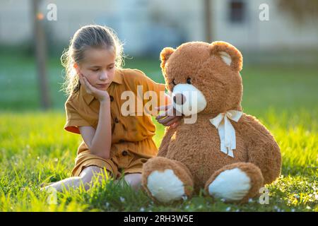 Triste bambina solitaria che passa del tempo insieme al suo amico orsacchiotto all'aperto nel soleggiato cortile. Solitudine all'età preadolescente Foto Stock