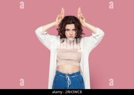 Ritratto di una donna arrabbiata aggressiva e attraente con i capelli ricci che indossa abiti casual con le corna del diavolo, pronta ad attaccare. Riprese in studio per interni isolate su sfondo rosa. Foto Stock