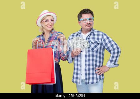 Un paio di amici, uomini e donne adulti in camicia informale a scacchi, in piedi insieme a una borsa della spesa e fan delle banconote da dollaro. Ripresa in studio in interni isolata, su sfondo giallo Foto Stock