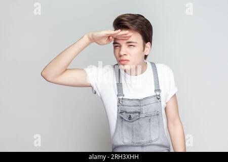 Ritratto di un giovane bruna serio che si trova in piedi tiene la mano vicino alla fronte, guarda lontano cerca qualcosa all'orizzonte, indossando tute in denim. Riprese in studio in interni isolate su sfondo grigio. Foto Stock