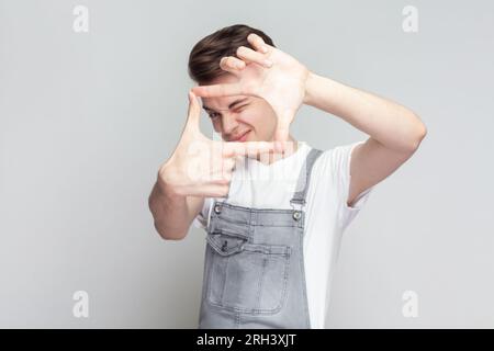 Cornice per dita gestanti. Ritratto di un giovane bruna che guarda la fotocamera e la cornice delle dita gestanti, indossando tute in denim. Riprese in studio in interni isolate su sfondo grigio. Foto Stock