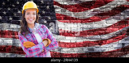 Appaltatore femminile che indossa elmetto giallo vuoto e guanti che sventolano la bandiera americana sullo sfondo del muro di mattoni. Foto Stock