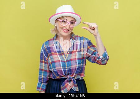 Ritratto di donna anziana che indossa camicia a scacchi, cappello e occhiali da vista mostra qualcosa di molto piccolo o minuscolo, discute i prezzi in negozio e sorride felicemente. Riprese in studio in interni isolate su sfondo giallo. Foto Stock