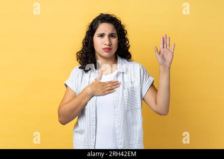 Ritratto di donna seria e responsabile attraente con capelli ondulati scuri che alzano il palmo per giurare, donna che giura di dire solo la verità. Riprese in studio in interni isolate su sfondo giallo. Foto Stock