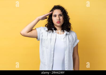 Ritratto di donna seria responsabile con capelli scuri ondulati che saluta il comandante, l'ordine di ascolto con espressione obbediente, guardando la fotocamera. Riprese in studio in interni isolate su sfondo giallo. Foto Stock