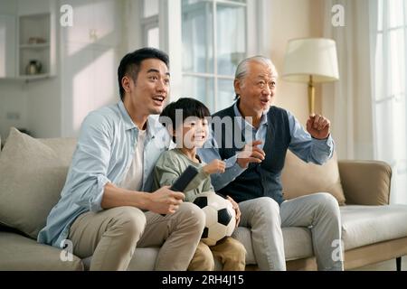 Figlio asiatico nonno seduto sul divano a casa a guardare la trasmissione in diretta della partita di calcio in TV insieme Foto Stock