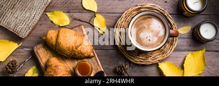 Concetto estetico autunnale. Tazza di cappuccino con croissant, candele, quadri e foglie gialle autunnali su fondo di legno, striscione. Foto Stock
