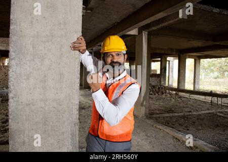 Ritratto di un giovane ingegnere civile indiano o architetto che indossa casco e giubbotto in piedi nel cantiere. Foto Stock