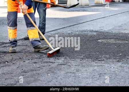 Gli operatori stradali rimuovono le macerie in eccesso con una spazzola e una pala sulla nuova superficie di asfalto intorno ai tombini fognari. Copia spazio. Foto Stock