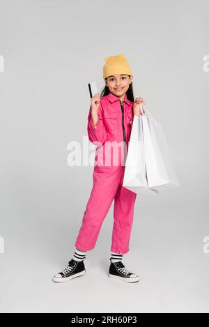 happy girl con berretto e outfit alla moda con carta di credito grigia, shopping per bambini, moda, gioia Foto Stock