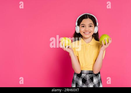 studentessa sorpresa con le cuffie wireless che reggono mela e zaino, sfondo rosa, studentessa Foto Stock