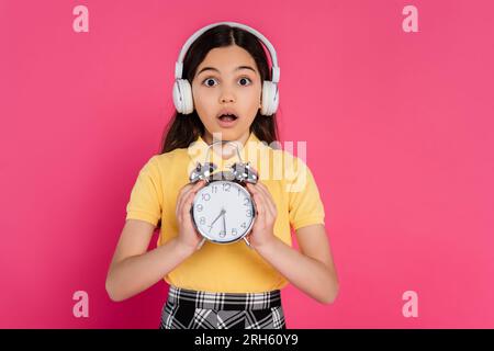 studentessa scioccata con le cuffie wireless che reggono una sveglia vintage isolata sulla vita studentesca rosa Foto Stock