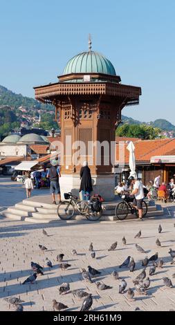 Ciclisti e piccioni vicino alla fontana Sebilj nel quartiere Baščaršija di Sarajevo, Bosnia ed Erzegovina, 14 agosto 2023. Foto Stock