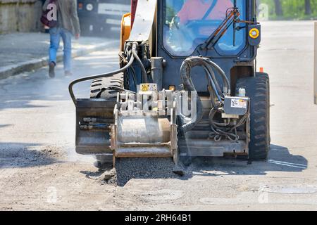 Un trattore per la costruzione di strade con una fresatrice montata ripara una sezione della strada rimuovendo uno strato di asfalto vecchio su una strada cittadina. Foto Stock