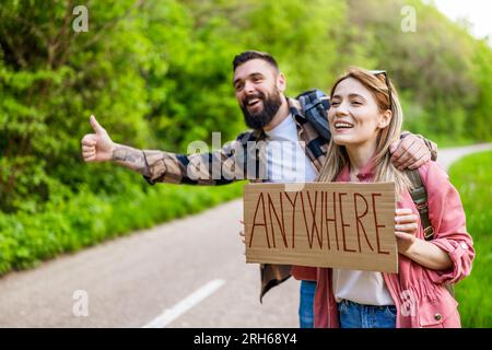 Felice coppia hitchhiking sulla strada cercando di fermare l'auto. Sono in cartoncino con iscrizione. Foto Stock