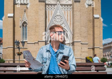 Uomo turista siede su una panchina nella piazza della città con mappa e cellulare e cerca l'hotel che ha prenotato tramite un'applicazione sul suo telefono viaggiatore maschio Foto Stock