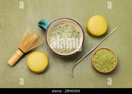 Una tazza di tè Matcha e tè Matcha in polvere in una ciotola. Frusta e cucchiaio di bambù. Torta al macaron. Sfondo verde Foto Stock