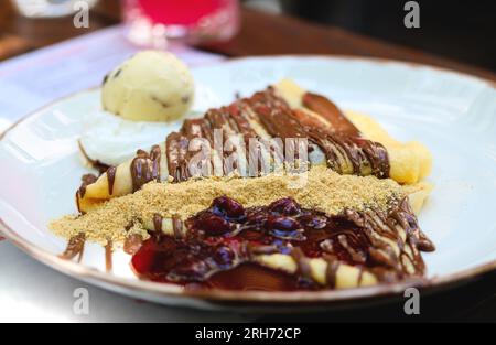 Crepes con crema al cioccolato. Pancake con pasta al cioccolato, nocciole, salsa di ciliegia e una paletta di gelato su un piatto bianco su un tavolo del ristorante. T Foto Stock