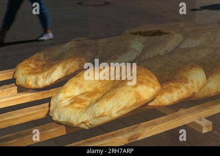 Il pane pita appena sfornato si trova su una griglia di legno in una panetteria. Vista dalla strada attraverso una vetrina di vetro che riflette le persone che passano. Foto Stock