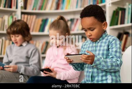 Diversi studenti scolastici che usano smartphone in biblioteca, bambini multirazziali che tengono telefoni cellulari, giocano Foto Stock