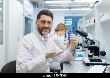 Ritratto di un giovane uomo sorridente che lavora in un laboratorio al microscopio, che tiene e mostra un pallone con una sostanza alla macchina fotografica. Una collega donna lavora accanto al tavolo. Foto Stock