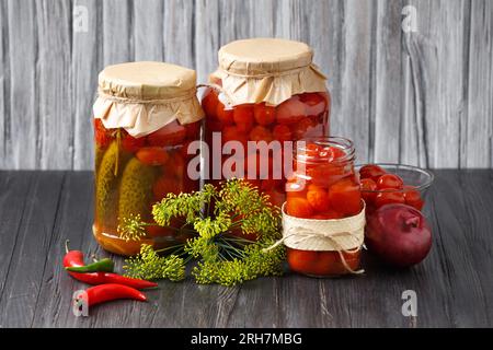 Pomodori ciliegini in scatola e cetrioli in vasetti e una ciotola, peperoncino piccante, cipolle su fondo di legno. Conserve fatte in casa, verdure sott'aceto. Foto Stock