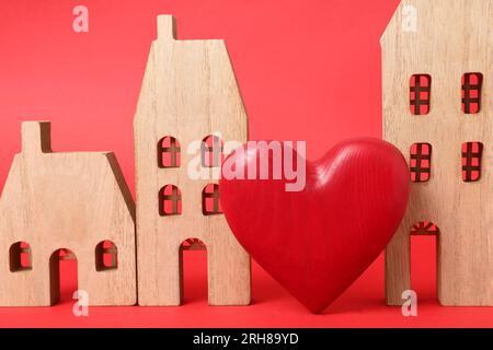 Concetto di relazione a distanza. Modelli di casa in legno e cuore decorativo su sfondo rosso, primo piano Foto Stock