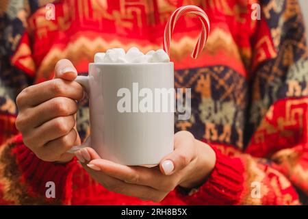 Donna con maglione rosso che regge una tazza bianca con cioccolata calda e primo piano di marshmallow. Modello di tazza di Natale Foto Stock