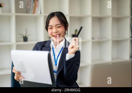 Una bella e allegra donna d'affari asiatica sta esaminando un rapporto d'affari nel suo ufficio. banchiere, avvocato, segretario, manager, contabile Foto Stock