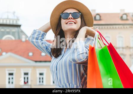 Ritratto di donna alla moda con cappello e occhiali da sole che tiene in mano colorate borse per la spesa mentre fa shopping in strada. Foto Stock