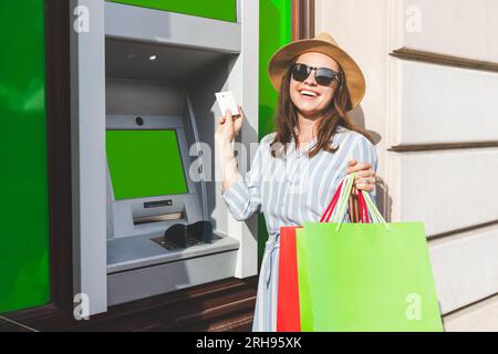 Una giovane donna alla moda si trova accanto al bancomat con in mano carta di credito e borse della spesa. Foto Stock