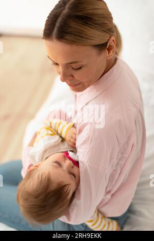 Madre amorevole che tiene la figlia addormentata sulle mani, lulling Kid, seduta sul letto, vista dall'alto, ripresa verticale Foto Stock