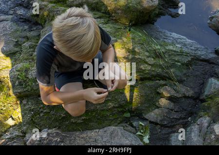 Un ragazzo seduto sulle rocce vicino al fiume equivale il gancio di un'esca. Pesca sportiva sul fiume in estate. Foto Stock