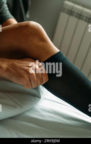 un uomo seduto sul letto indossa un calzino a compressione nero, con la luce del giorno Foto Stock