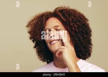 Il giovane con i capelli ricci dello zenzero entra nella sua routine di cura della pelle, applicando una crema di bellezza sul suo viso lentigginato. Uomo con caratteristiche di bellezza uniche coccola h Foto Stock
