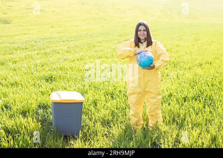 Una donna in tuta protettiva gialla si trova al centro di un campo verde e tiene un globo nelle sue mani, accanto a un cestino di rifiuti. Cura del pianeta, salva Foto Stock