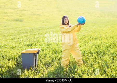 Una donna in tuta protettiva gialla si trova al centro di un campo verde e tiene un globo nelle sue mani, accanto a un cestino di rifiuti. Cura del pianeta, salva Foto Stock
