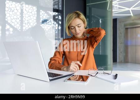 Donna d'affari stanca sul posto di lavoro, lavora a lungo in ufficio, donna malata soffre di gravi dolori al collo e lavora al computer portatile mentre è seduta. Foto Stock